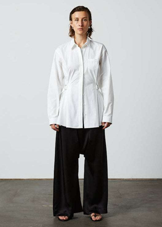 INTERNATIONAL CITIZEN.  Style: Artist Shirt. Front full body view. Style has an oversized shirt with a pen pocket at the chest, open underarms for breathability, and back belt ties that adjust at the front with silver rings. The shirt is made of Italian organic Cotton in White Quartz Spiritually Infused, Sustainable Luxury Fashion in Berlin.