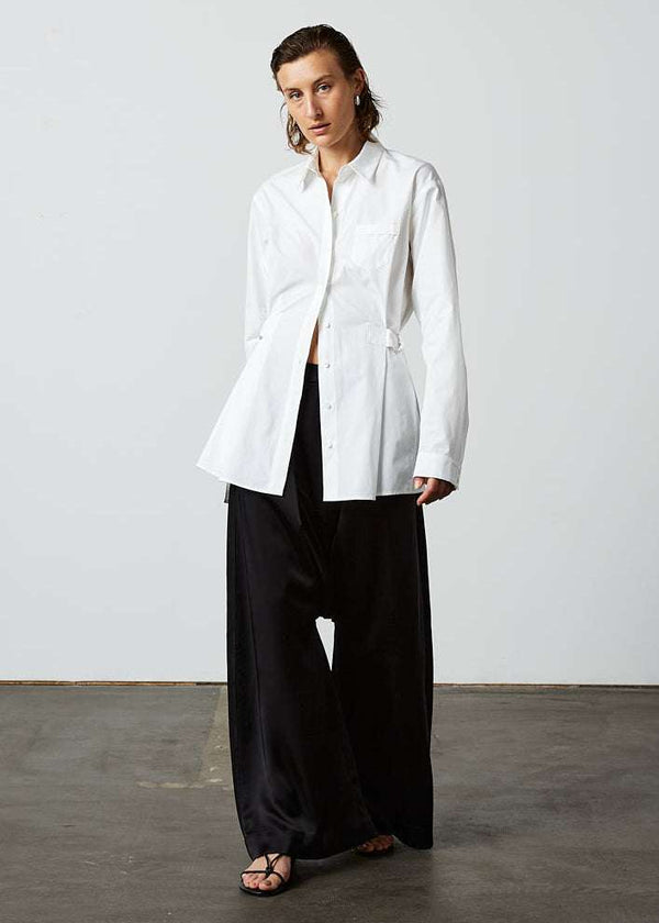 INTERNATIONAL CITIZEN.  Style: Artist Shirt. Front view worn slightly un-buttoned. Style has an oversized shirt with a pen pocket at the chest, open underarms for breathability, and back belt ties that adjust at the front with silver rings. The shirt is made of Italian organic Cotton in White Quartz Spiritually Infused, Sustainable Luxury Fashion in Berlin.
