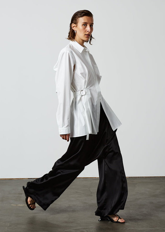 INTERNATIONAL CITIZEN.  Style: Artist Shirt. Side view. Style has an oversized shirt with a pen pocket at the chest, open underarms for breathability, and back belt ties that adjust at the front with silver rings. The shirt is made of Italian organic Cotton in White Quartz Spiritually Infused, Sustainable Luxury Fashion in Berlin.