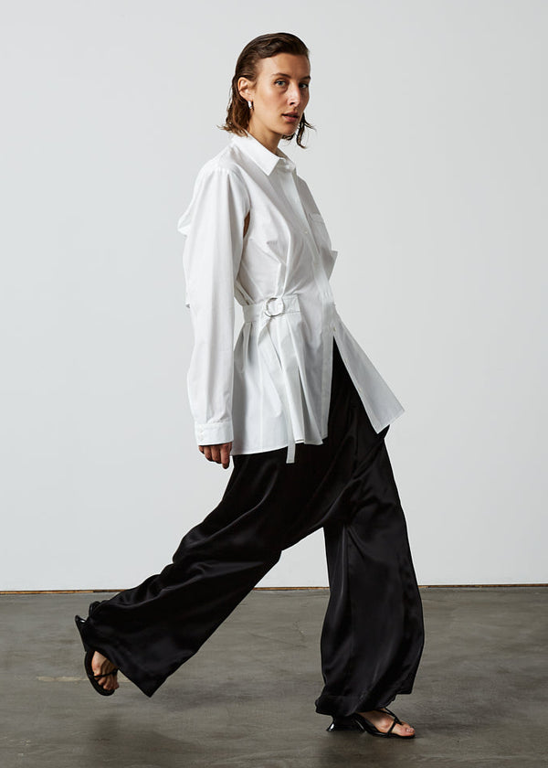 INTERNATIONAL CITIZEN.  Style: Artist Shirt. Side view. Style has an oversized shirt with a pen pocket at the chest, open underarms for breathability, and back belt ties that adjust at the front with silver rings. The shirt is made of Italian organic Cotton in White Quartz Spiritually Infused, Sustainable Luxury Fashion in Berlin.