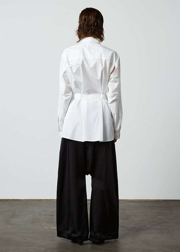 INTERNATIONAL CITIZEN.  Style: Artist Shirt. Back full body view. Style has an oversized shirt with a pen pocket at the chest, open underarms for breathability, and back belt ties that adjust at the front with silver rings. The shirt is made of Italian organic Cotton in White Quartz Spiritually Infused, Sustainable Luxury Fashion in Berlin.