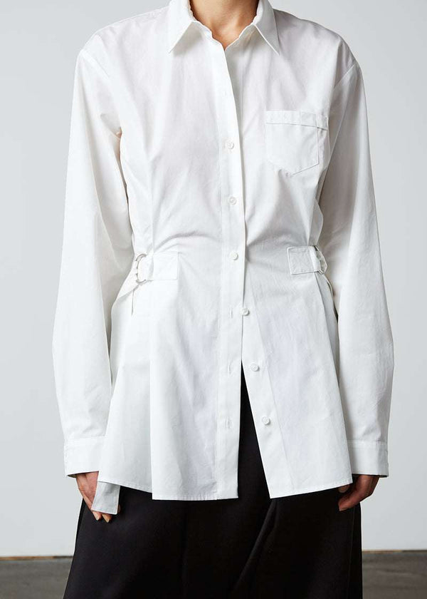 INTERNATIONAL CITIZEN.  Style: Artist Shirt. Close up front photo. Style has an oversized shirt with a pen pocket at the chest, open underarms for breathability, and back belt ties that adjust at the front with silver rings. The shirt is made of Italian organic Cotton in White Quartz Spiritually Infused, Sustainable Luxury Fashion in Berlin.