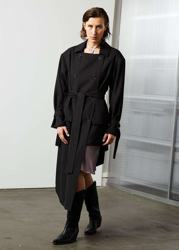 INTERNATIONAL CITIZEN.  Style: The Eclipse Jacket. A unisex, asymmetrical along the hemline and oversized with soft shoulder pads for collapsed structure. It is belted, double breasted, has extra-large pockets, and adjustable sleeve straps with silver rings.  Front view. Made of Italian RWS Wool, Obsidian Black. Spiritually Infused, Sustainable Luxury Fashion in Berlin.