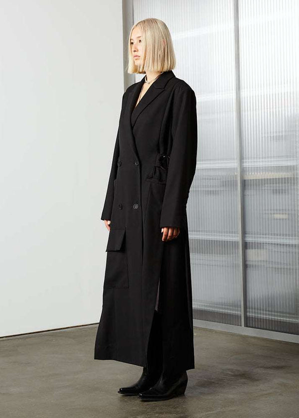 INTERNATIONAL CITIZEN.  Style: The M23 Duster Coat is double breasted with button closures, and features high slits at the sides and back, an extra large low positioned front pocket. Unisex Item. Side front view. Made of Italian Wool RWS, Obsidian Black. Spiritually Infused, Sustainable Luxury Fashion in Berlin.