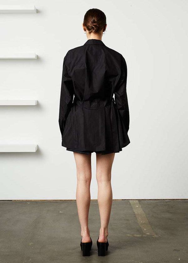 INTERNATIONAL CITIZEN.  Style: Artist Shirt. Back full body view. Style has an oversized shirt with a pen pocket at the chest, open underarms for breathability, and back belt ties that adjust at the front with silver rings. The shirt is made of Italian organic Cotton in Obsidian Black. Spiritually Infused, Sustainable Luxury Fashion in Berlin.