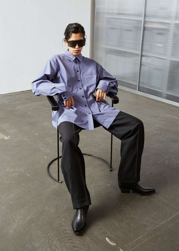INTERNATIONAL CITIZEN.  Style: Artist Shirt. Model sitting in a chair. Style has an oversized shirt with a pen pocket at the chest. The shirt is made of Italian organic Cotton in Dreamstate Purple. Spiritually Infused, Sustainable Luxury Fashion in Berlin.
