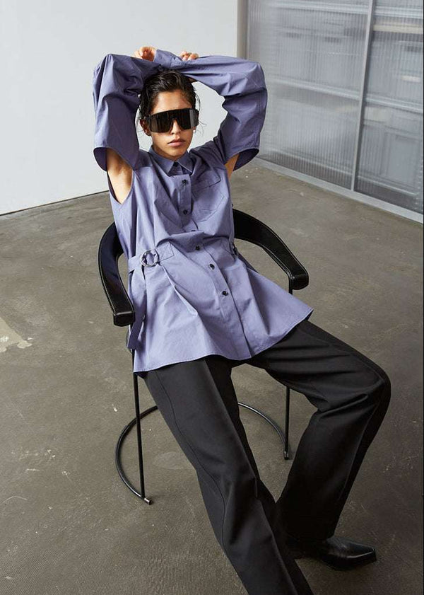 INTERNATIONAL CITIZEN.  Style: Artist Shirt. Style has an oversized shirt with a pen pocket at the chest, open underarms for breathability, and back belt ties that adjust at the front with silver rings. The shirt are made of Italian organic Cotton in Dreamstate Purple. Model sitting back in the chair with her hands up. Spiritually Infused, Sustainable Luxury Fashion in Berlin.