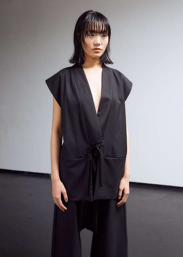 INTERNATIONAL CITIZEN.  Style: The Kimono Vest has a longer front body with inseam pockets and short back body. There are front ties and inseam belt slits for multiple ways to belt it. Unisex Item. Female front photo. Made of Italian Wool, certified (RWS) Responsible Wool Standard, Obsidian Black. Spiritually Infused, Sustainable Luxury Fashion in Berlin.
