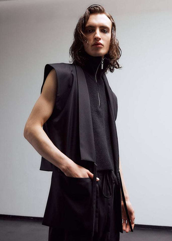INTERNATIONAL CITIZEN.  Style: The Kimono Vest has a longer front body with inseam pockets and short back body. Unisex Item. Side front male model. Made of Italian Wool, certified (RWS) Responsible Wool Standard, Obsidian Black. Spiritually Infused, Sustainable Luxury Fashion in Berlin.