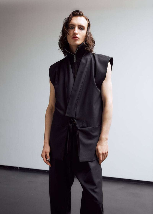 INTERNATIONAL CITIZEN.  Style: The Kimono Vest has a longer front body with inseam pockets and short back body. There are front ties and inseam belt slits for multiple ways to belt it. Unisex Item. Male front photo. Made of Italian Wool, certified (RWS) Responsible Wool Standard, Obsidian Black. Spiritually Infused, Sustainable Luxury Fashion in Berlin.