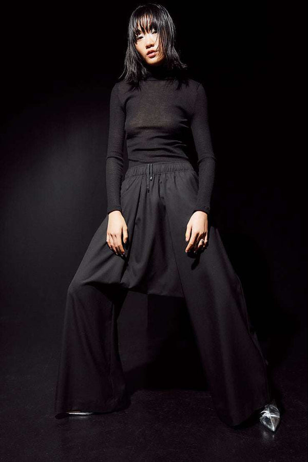 INTERNATIONAL CITIZEN.  Style: The Lanna Pant have a dropped crotch with excessive volume eased into an elastic waistband. Unisex Item. Front female photo with movement. Made of Italian Wool, certified (RWS) Responsible Wool Standard, Obsidian Black. Spiritually Infused, Sustainable Luxury Fashion in Berlin.

