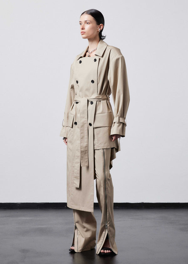 INTERNATIONAL CITIZEN.  Style: The Eclipse Jacket. A unisex, asymmetrical along the hemline and oversized with soft shoulder pads for collapsed structure. It is half lined, belted, double breasted, has extra-large pockets, and adjustable sleeve straps with silver rings. Side front view.  Made of Italian Organic Cotton, Dune. Spiritually Infused, Sustainable Luxury Fashion in Berlin.