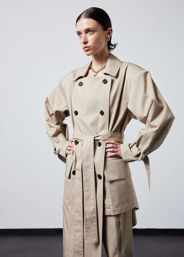 INTERNATIONAL CITIZEN.  Style: The Eclipse Jacket. A unisex, asymmetrical along the hemline and oversized with soft shoulder pads for collapsed structure. It is  belted, double breasted, has extra-large pockets, and adjustable sleeve straps with silver rings. Close up side front view.  Made of Italian Organic Cotton, Dune. Spiritually Infused, Sustainable Luxury Fashion in Berlin.