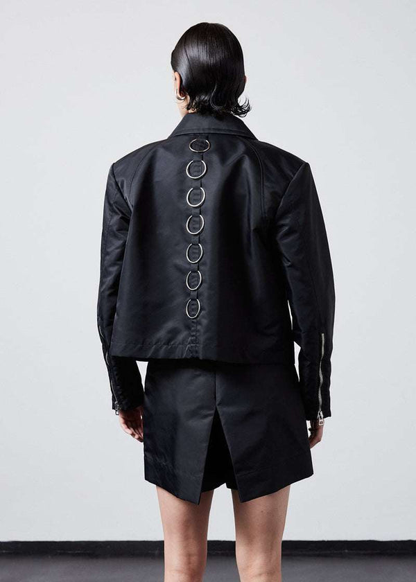 INTERNATIONAL CITIZEN.  Style: Ajna Jacket. Back view of a black boxy jacket with sporty tailoring, featuring square shoulders, tapered sleeves, and zipper closures. The jacket features 7-rings down the back body. The jacket is made from of Italian water repellent, post-consumer recycled Nylon Gaberdine. Spiritually Infused, Sustainable Luxury Fashion in Berlin.