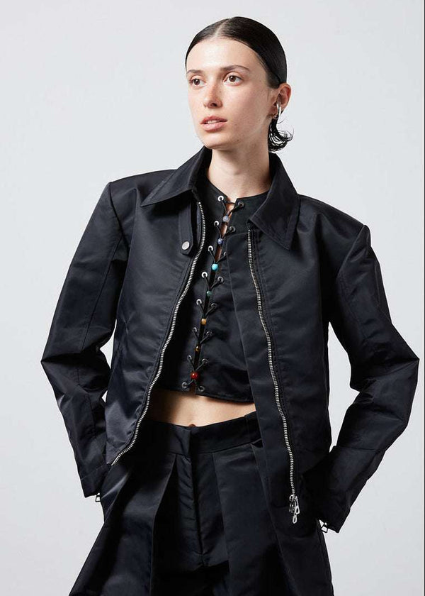 INTERNATIONAL CITIZEN.  Style: Ajna Jacket. Front view of a black boxy jacket worn open with sporty tailoring, featuring square shoulders, tapered sleeves, and zipper closures. The jacket has a snap tab at the collar neckline. The jacket is made from of Italian water repellent, post-consumer recycled Nylon Gaberdine. Spiritually Infused, Sustainable Luxury Fashion in Berlin.