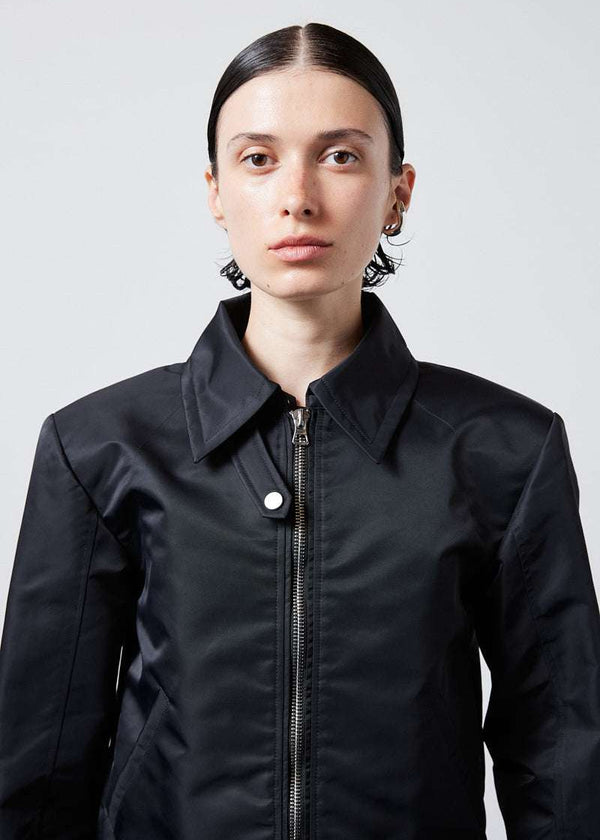 INTERNATIONAL CITIZEN.  Style: Ajna Jacket. Close-up photo of a black boxy jacket with sporty tailoring, featuring square shoulders, tapered sleeves, and zipper closures. The jacket has a snap tab at the collar neckline. The jacket is made from of Italian water repellent, post-consumer recycled Nylon Gaberdine. Spiritually Infused, Sustainable Luxury Fashion in Berlin.