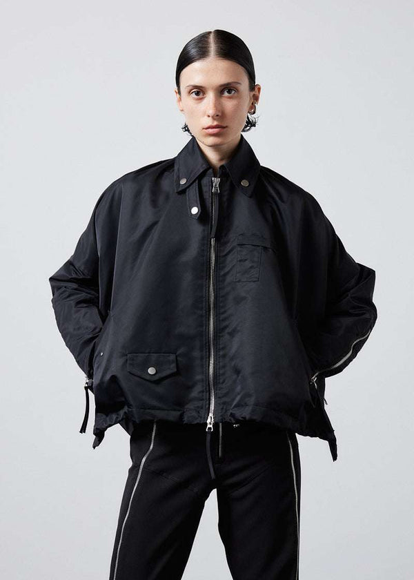 INTERNATIONAL CITIZEN.  Style: The Cube Coat, is a 3D cube tilted on its axes to create a boxy minimal front body and dramatic drape in the back body. It has zipper closures, and a drawcord at the front hemline and a utility pen pocket on the chest. Unisex style. Front photo. Made of Italian post-consumer recycled Nylon, Obsidian Black. Spiritually Infused, Sustainable Luxury Fashion in Berlin.