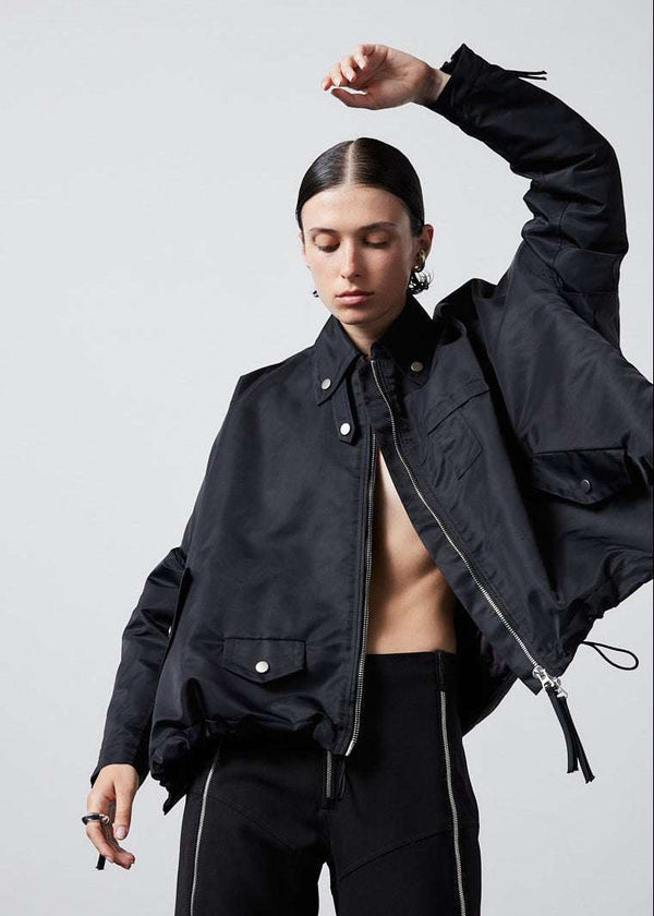INTERNATIONAL CITIZEN.  Style: The Cube Coat, is a 3D cube tilted on its axes to create a boxy minimal front body and has zipper closures, and a drawcord at the front hemline and a utility pen pocket on the chest. Unisex style. Close up front view. Made of Italian post-consumer recycled Nylon, Obsidian Black. Spiritually Infused, Sustainable Luxury Fashion in Berlin.