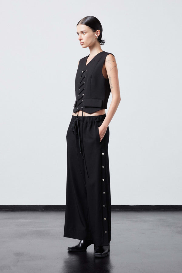 INTERNATIONAL CITIZEN.  Style: The Track Trouser has a smooth back waistband and elastic front with a drawcord tunnel at the front. Silver metal snaps down the side and cropped ankle length. Unisex item. Made of Italian Wool RWS, Obsidian Black. Spiritually Infused, Sustainable Luxury Fashion in Berlin. Full body side front photo.