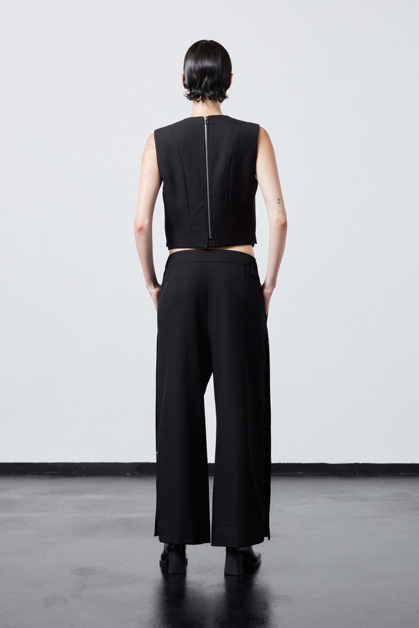 INTERNATIONAL CITIZEN.  Style: The Track Trouser has a smooth back waistband and elastic front with a drawcord tunnel at the front. Silver metal snaps down the side and cropped ankle length. Unisex item. Made of Italian Wool RWS, Obsidian Black. Spiritually Infused, Sustainable Luxury Fashion in Berlin. Full back body photo.