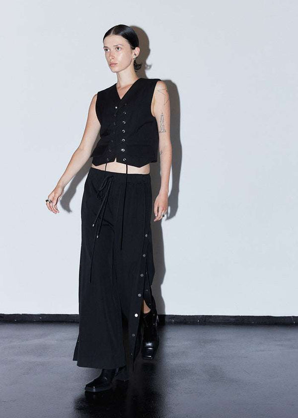 INTERNATIONAL CITIZEN.  Style: The Track Trouser has a smooth back waistband and elastic front with a drawcord tunnel at the front. Silver metal snaps down the side and cropped ankle length. Unisex item. Made of Italian Wool RWS, Obsidian Black. Spiritually Infused, Sustainable Luxury Fashion in Berlin. Walking front photo.