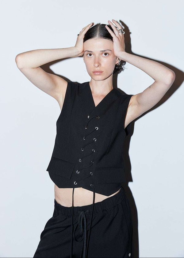 INTERNATIONAL CITIZEN.  Style: The Stitch Vest features a high neckline with overlapping front closure held with an eyelet stitch lacing detail. The back has an exposed zipper for easy wearability and front flap welt pockets.Made of Italian Wool RWS, Obsidian Black. Spiritually Infused, Sustainable Luxury Fashion in Berlin. Front photo.