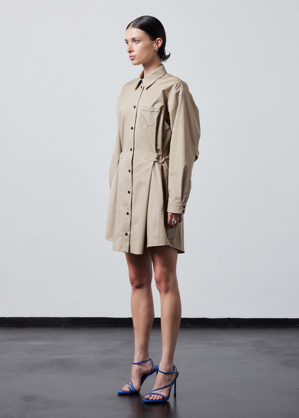 INTERNATIONAL CITIZEN.  Style: Artist Shirt Dress. Side front view. Style has an oversized shirt with a pen pocket at the chest, open underarms for breathability, and back belt ties that adjust at the front with silver rings. The shirt are made of Italian organic Cotton Twill in Dune. Spiritually Infused, Sustainable Luxury Fashion in Berlin.