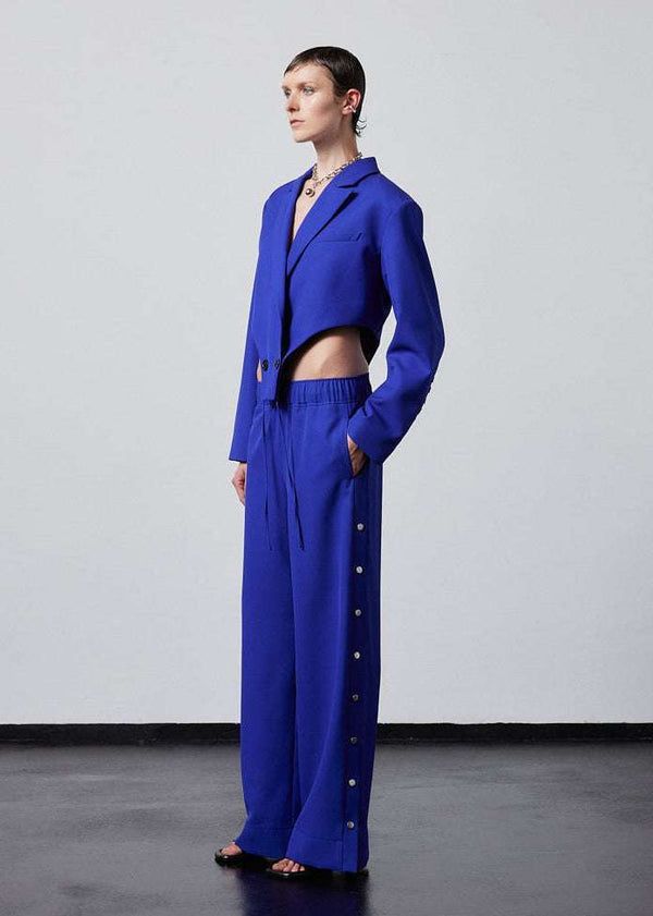 INTERNATIONAL CITIZEN.  Style: The Track Trouser has a smooth back waistband and elastic front with a drawcord tunnel at the front. Silver metal snaps down the side and cropped ankle length. Unisex item. Made of Italian Wool RWS, Lapis Lazuli Blue. Spiritually Infused, Sustainable Luxury Fashion in Berlin. Side front view.