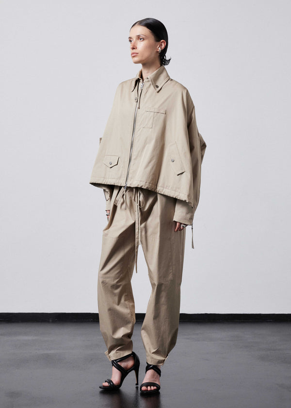 INTERNATIONAL CITIZEN.  Style: The Cube Coat, is a 3D cube tilted on its axes to create a boxy minimal front body and dramatic drape in the back body. It has zipper closures, and a drawcord at the front hemline and a utility pen pocket on the chest. Unisex style. Side front view. Made of Italian Organic Cotton Twill, Dune. Spiritually Infused, Sustainable Luxury Fashion in Berlin.