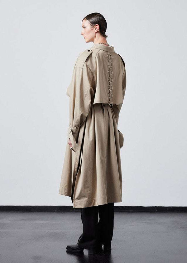 INTERNATIONAL CITIZEN.  Style: The Equinox Trench Coat, is voluminous, it features classic trench coat detailing and optional 7-Chakra ring detail at the back storm flap. Unisex item.  Side back view. Made of Italian Organic Cotton, Dune. Spiritually Infused, Sustainable Luxury Fashion in Berlin.
