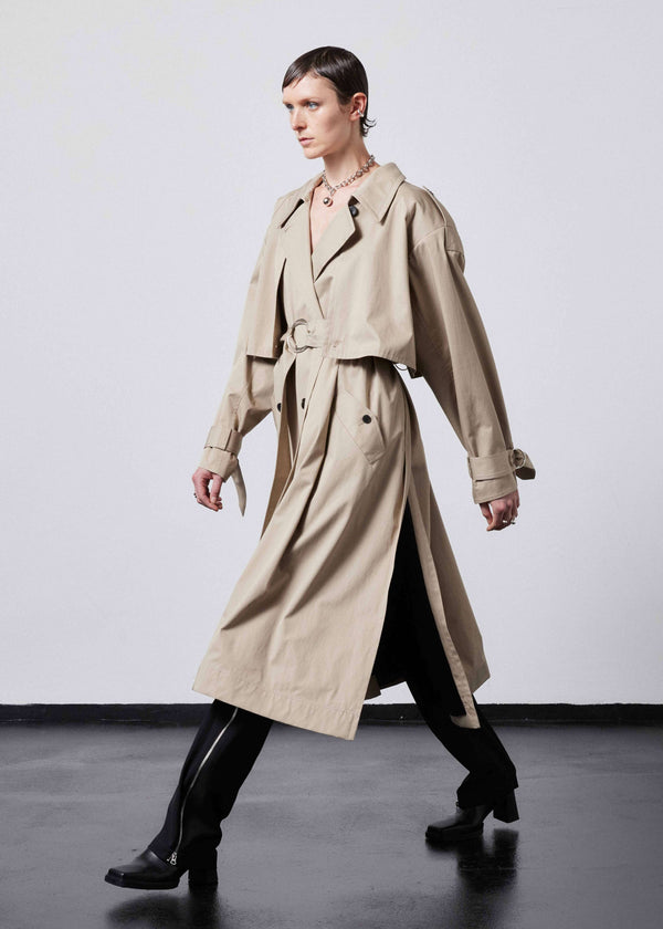 INTERNATIONAL CITIZEN.  Style: The Equinox Trench Coat, is voluminous and it features classic trench coat detailing, overlapping sides seams at the underarm, and a storm flap. Unisex item. Profile view walking. Made of Italian Organic Cotton, Dune. Spiritually Infused, Sustainable Luxury Fashion in Berlin.