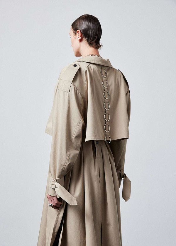 INTERNATIONAL CITIZEN.  Style: The Equinox Trench Coat, is voluminous, it features classic trench coat detailing, overlapping sides seams at the underarm, removable shoulder pads and optional 7-Chakra ring detail at the back storm flap. Unisex item.  Close up side back view. Made of Italian Organic Cotton, Dune. Spiritually Infused, Sustainable Luxury Fashion in Berlin.