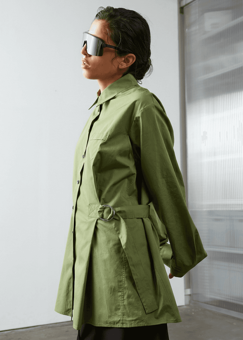 INTERNATIONAL CITIZEN.  Style: Artist Shirt Dress. Side view. The style has an oversized shirt with a pen pocket at the chest and back belt ties that adjust at the front with silver rings. The shirt are made of Italian organic Cotton Twill in Nephrite Jade. Spiritually Infused, Sustainable Luxury Fashion in Berlin.