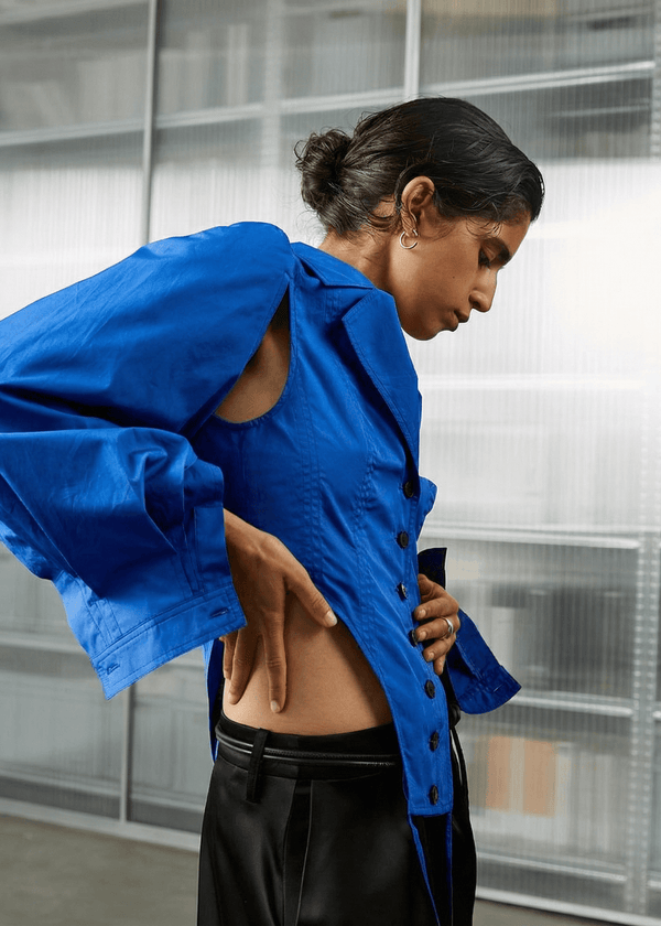 INTERNATIONAL CITIZEN.  Style: A French Cut Button Bodysuit with tie closures. It features exaggerated sleeve volume pleated at the cuffs, with an open under arm for breathability. Side front view. Made of Italian Organic Cotton, Lapis lazuli Blue. Spiritually Infused, Sustainable Luxury Fashion in Berlin.