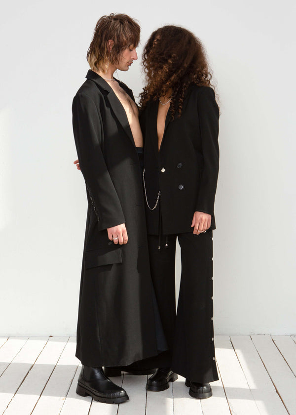 INTERNATIONAL CITIZEN.  Style: The Heart Opener Jacket features slim sleeves cut on the bias. Photo with male model beside women in the jacket. Made of Italian Wool, certified (RWS) Responsible Wool Standard, Obsidian Black. Spiritually Infused, Sustainable Luxury Fashion in Berlin.