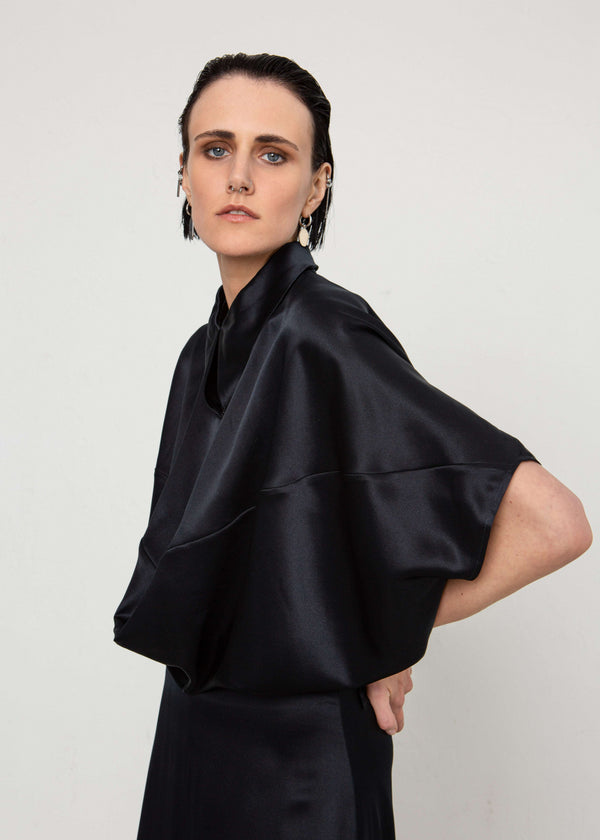 INTERNATIONAL CITIZEN.  Style: Prism Top, using sacred geometry of a 3-dimensional prism falling over the body in a soft cowl drape with an origami folded collar at the front placket. Made of Organic Silk, Obsidian Black. Spiritually Infused, Sustainable Luxury Fashion in Berlin. Side front photo.