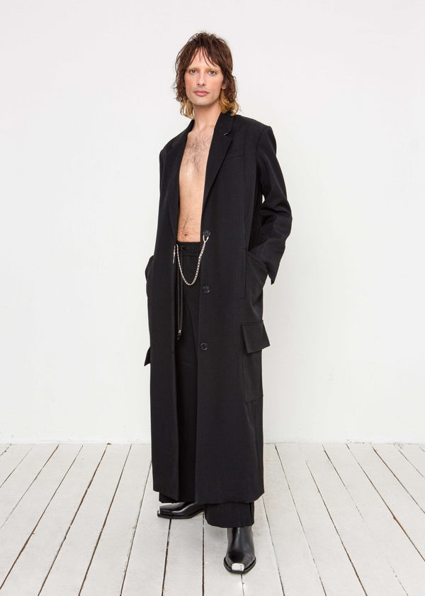 INTERNATIONAL CITIZEN.  Style: Duster Coat. A unisex coat that features traditional men's tailoring construction details. Male model front view. The coat is made of Italian RWS Wool in Obsidian Black. Spiritually Infused, Sustainable Luxury Fashion in Berlin.