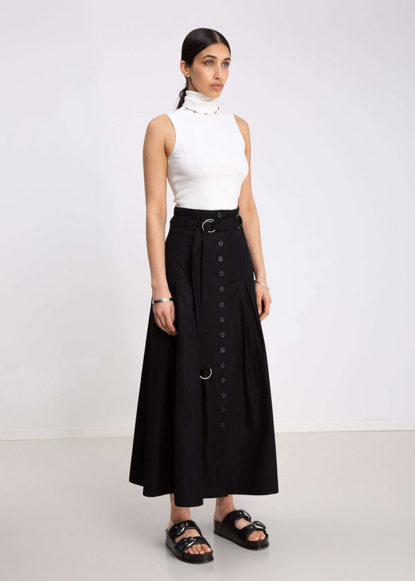 INTERNATIONAL CITIZEN.  Style: Button Skirt.  Side front view. A Button Skirt with box pleats below the hips. Detachable belt with silver ring details for closure. The skirt is made of Italian organic Cotton in Obsidian Black. Spiritually Infused, Sustainable Luxury Fashion in Berlin.