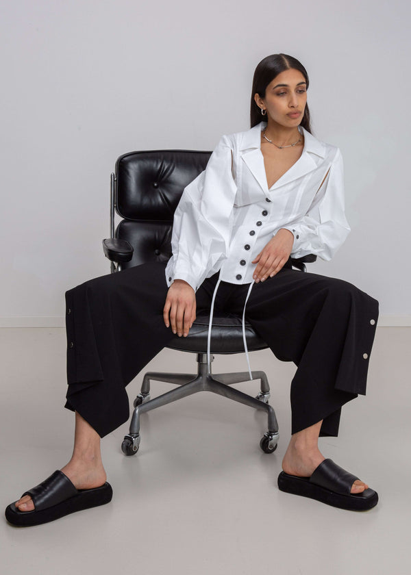 INTERNATIONAL CITIZEN.  Style: A French Cut Button Bodysuit with tie closures. It features exaggerated sleeve volume pleated at the cuffs. Seated front photo. Made of Italian Organic Cotton, White Quartz. Spiritually Infused, Sustainable Luxury Fashion in Berlin.