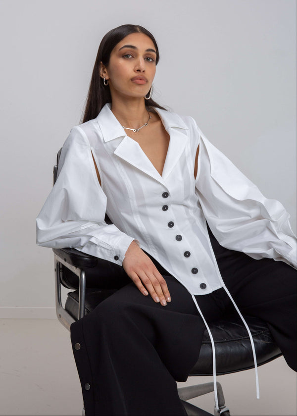 INTERNATIONAL CITIZEN.  Style: A French Cut Button Bodysuit with tie closures. It features exaggerated sleeve volume pleated at the cuffs, with an open under arm for breathability. Seated photo of the front. Made of Italian Organic Cotton, White Quartz. Spiritually Infused, Sustainable Luxury Fashion in Berlin.